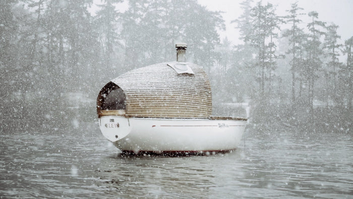 A unique sailboat with a custom cabin and chimney made from wood floats on the water with trees in the background. It is a winter scene with snow falling in large snowflakes. video production videography videographer victoria bc