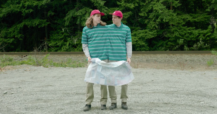 Adult conjoined twins face the camera wearing a custom-made collared shirt. They are both wearing matching red hats and are both holding a paper map. Green forest foliage in the background. video production videography videographer victoria bc