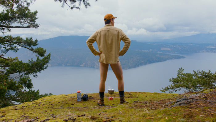 A man without pants standing atop the edge of a lookout. there is a large body of water and mountains in the distance. The man has a camping stove and water bottle. video production videography videographer victoria bc