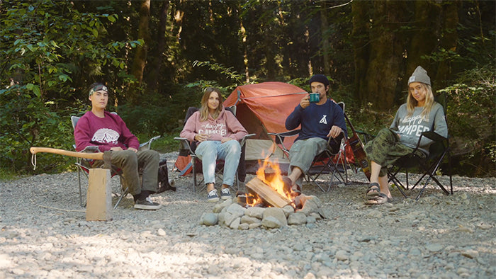 Four friends sit around a campfire at a camp ground. There is an axe placed atop a stump on the left and an orange tent behind the four people. The background is a dark forest. video production videography videographer victoria bc